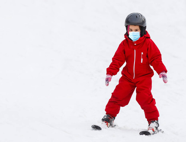 Πορτρέτο ενός μικρού κοριτσιού σκιέρ με ιατρική μάσκα κατά τη διάρκεια του COVID-19 coronavirus σε ένα χιονισμένο βουνό σε ένα χιονοδρομικό κέντρο - Φωτογραφία, εικόνα