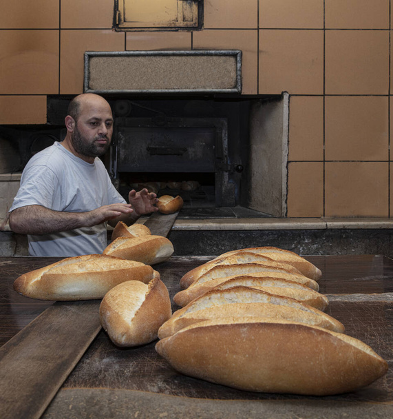 Fırıncı. Genç, yakışıklı bir fırın işçisi ekmek arka planında, sobadan tahta bir kürekle ekmek alıyor. Fırın ürünlerinin endüstriyel üretimi. Fırıncının özel giysileri içinde bir adam. Fırın - Fotoğraf, Görsel