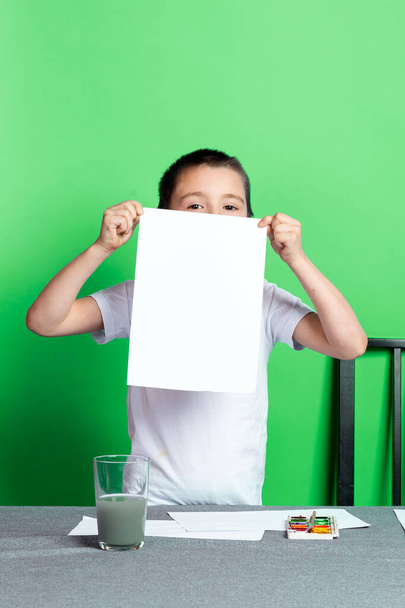 Creativiteit van kinderen. Mock-up, een jongen toont een blanco A4-vel als zijn tekening op een groene achtergrond - Foto, afbeelding