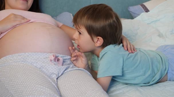 Portré kisfiúról pizsamában, aki terhes anyával fekszik az ágyban, és a meg nem született babával beszél a nagy hasában. Szerető és gondoskodó gyermekek - Felvétel, videó