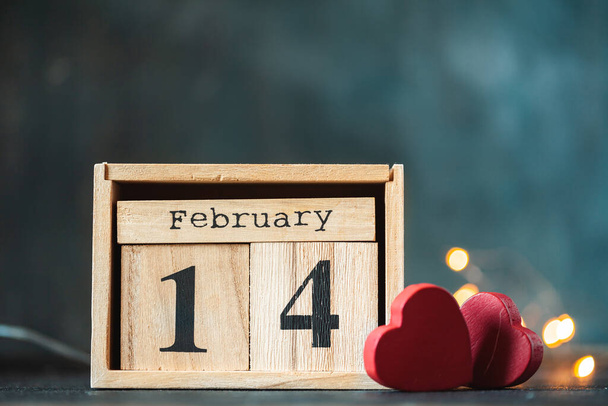 Wooden calendar of blocks with the date of February 14 on the blurred dark concrete background. two red hearts with bokeh lights behind. Happy Valentine's day. - Photo, Image
