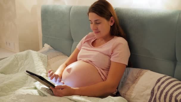 Portrait of smiling pregnant woman in pajamas lying in bed at night and browsing internet or social media on tablet computer - Footage, Video