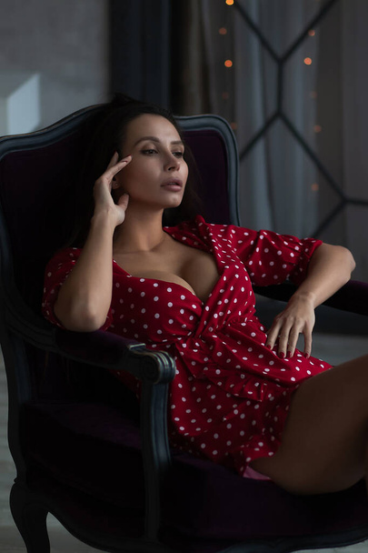 Sensual young female with long dark hair in stylish dress with decollete relaxing in comfortable armchair and looking away - Foto, imagen