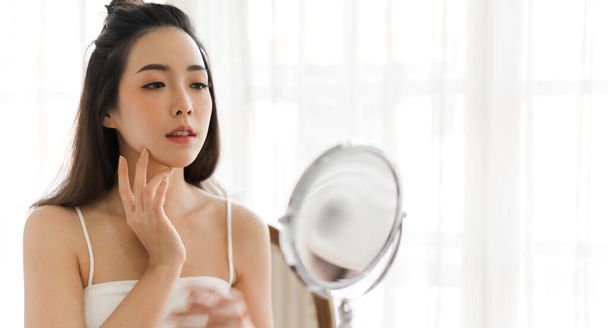 Smiling of young beautiful pretty asian woman clean fresh healthy white skin looking at mirror.girl touching on her face with hand and applying cream at home.spa and beauty concept - Photo, Image