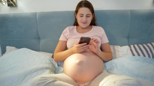 Giovane donna incinta in pigiama sdraiata a letto e utilizzando lo smartphone. Donna incinta con telefono sdraiata a letto la notte - Filmati, video