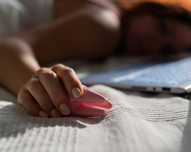 A woman lies in bed holding a clitoral vibrator and watching porn on a laptop. The girl has sex online - Foto, Imagen