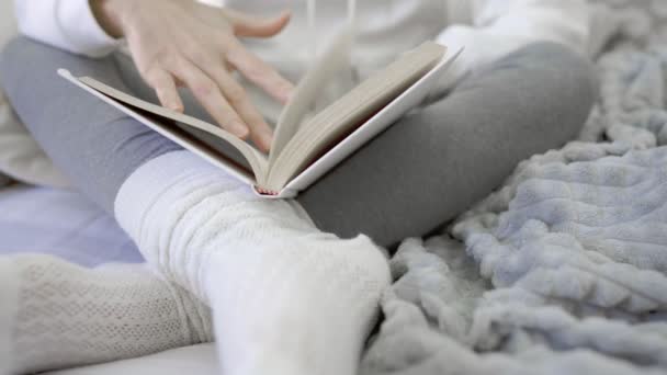 Vista inferior de una joven irreconocible sentada en el sofá con las piernas cruzadas leyendo un libro apoyado en calcetines de lana blanca siguiendo con el dedo las líneas de texto en colores blancos y grises de invierno - Imágenes, Vídeo