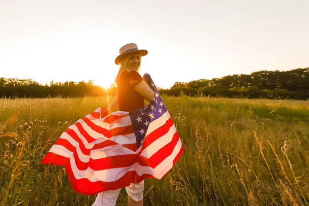 Beautiful young girl holding an American flag in the wind in a field of rye. Summer landscape against the blue sky. Horizontal orientation. - Photo, Image