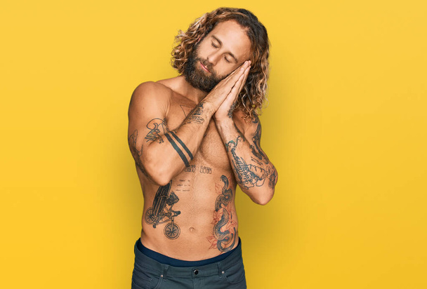 Handsome man with beard and long hair standing shirtless showing tattoos sleeping tired dreaming and posing with hands together while smiling with closed eyes.  - Photo, Image
