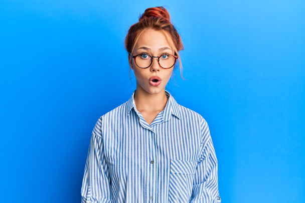 Young beautiful redhead woman wearing casual clothes and glasses over blue background scared and amazed with open mouth for surprise, disbelief face  - Photo, Image
