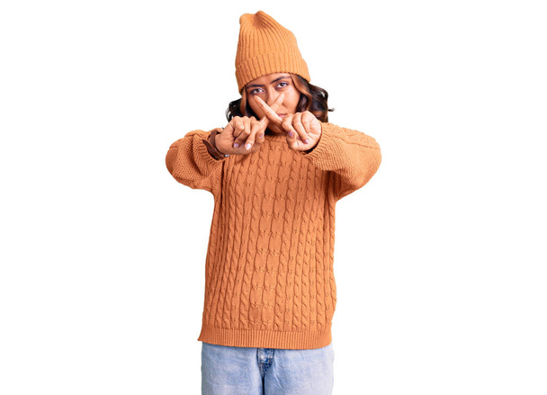 Young beautiful mixed race woman wearing wool sweater and winter hat rejection expression crossing fingers doing negative sign  - Foto, afbeelding