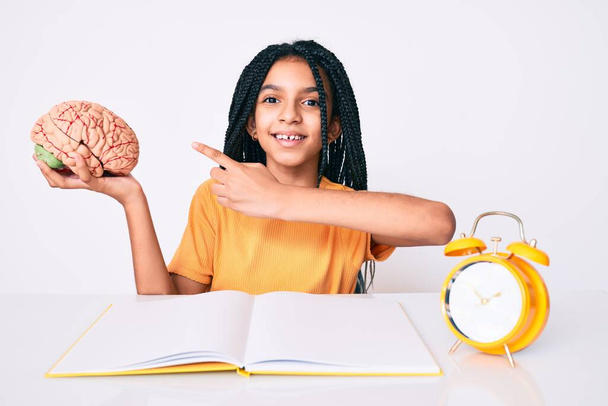 Junge afrikanisch-amerikanische Mädchen mit Zöpfen, die das Gehirn halten, während sie für die Schule lernen, lächeln glücklich und zeigen mit Hand und Finger  - Foto, Bild