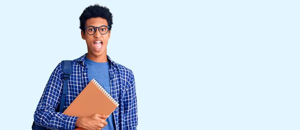 Young african american man wearing student backpack holding book sticking tongue out happy with funny expression. emotion concept.  - Zdjęcie, obraz