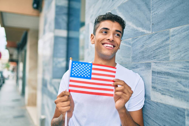 Jovem latino sorrindo feliz segurando bandeira americana encostada na parede da cidade. - Foto, Imagem