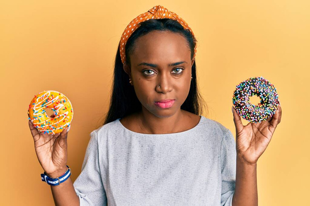 Νεαρή Αφρικανή γυναίκα που κρατά νόστιμα πολύχρωμα ντόνατς χαλαρά με σοβαρή έκφραση στο πρόσωπο. απλή και φυσική κοιτάζοντας την κάμερα.  - Φωτογραφία, εικόνα