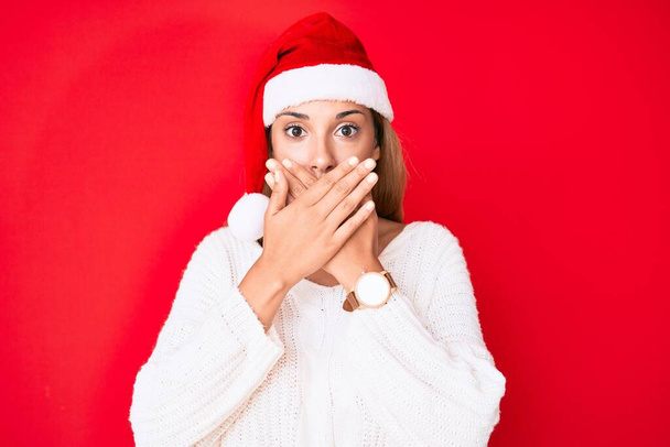 クリスマスの帽子をかぶっている若いブルネットの女性は間違いのために手で口を覆いショックを受けた。秘密の概念.  - 写真・画像