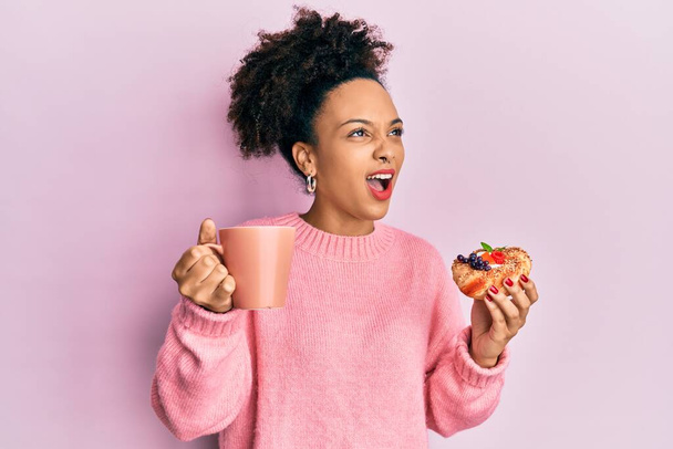 Young african american girl drinking a cup of coffee and eating sweet angry and mad screaming frustrated and furious, shouting with anger looking up.  - Photo, image