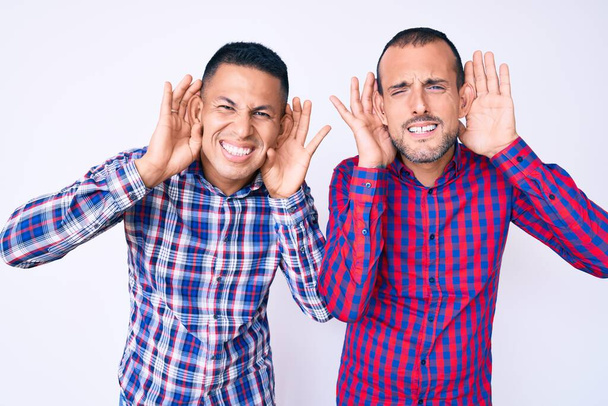 Молодые геи двое мужчин в повседневной одежде пытаются услышать жесты обеих рук, любопытные для сплетен. проблема со слухом, глухой  - Фото, изображение