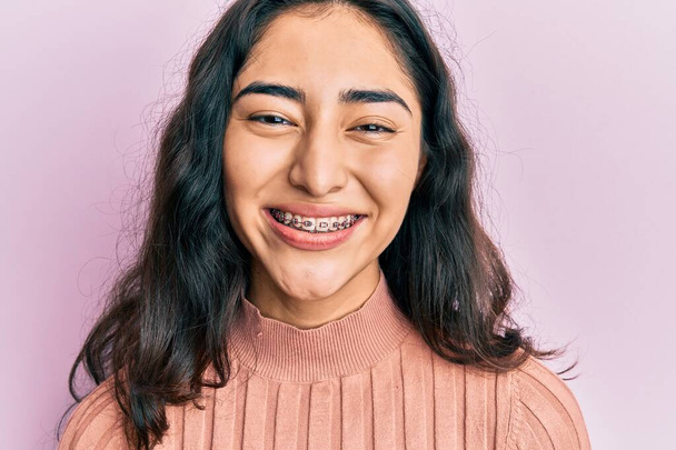 Латиноамериканская девочка-подросток с зубными скобками, показывающими ортодонтические скобки, выглядит позитивно и счастливо стоящей и улыбающейся с уверенной улыбкой, показывающей зубы  - Фото, изображение
