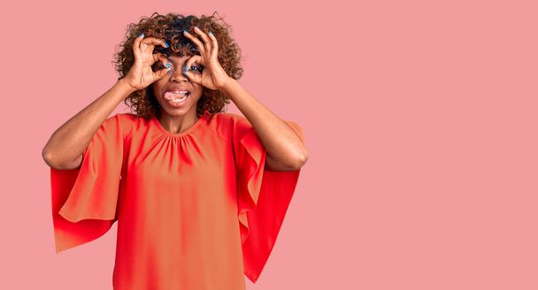 Joven mujer afroamericana vestida con ropa casual haciendo un buen gesto como binoculares sacando la lengua, ojos mirando a través de los dedos. expresión loca.  - Foto, imagen
