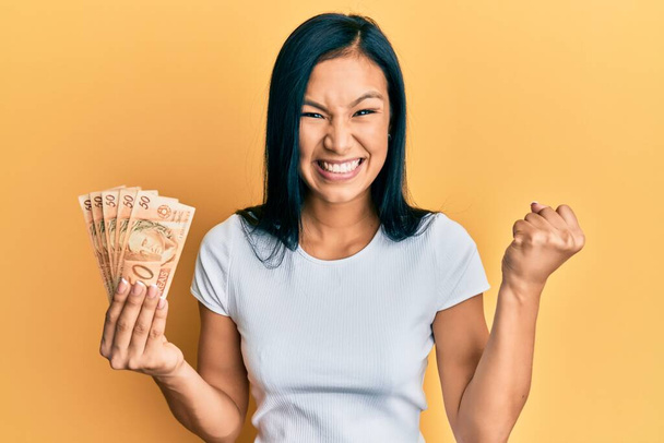 Beautiful hispanic woman holding 50 brazilian real banknotes screaming proud, celebrating victory and success very excited with raised arm  - Photo, Image