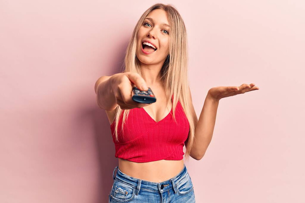 Young blonde woman holding television remote control celebrating achievement with happy smile and winner expression with raised hand  - Photo, Image