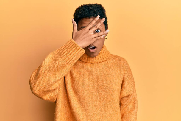 Mladá afroameričanka v neformálním oblečení vykukuje v šoku a zakrývá obličej a oči rukou, dívá se prsty s rozpačitým výrazem.  - Fotografie, Obrázek
