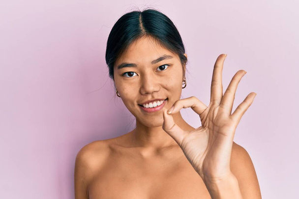 Young chinese woman standing topless showing skin smiling positive doing ok sign with hand and fingers. successful expression.  - Fotografie, Obrázek