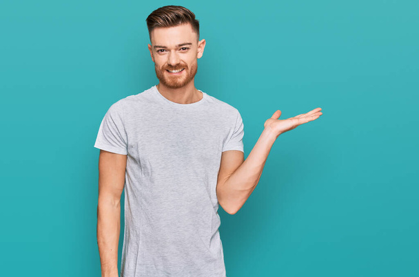 Young redhead man wearing casual grey t shirt smiling cheerful presenting and pointing with palm of hand looking at the camera.  - Foto, Imagem