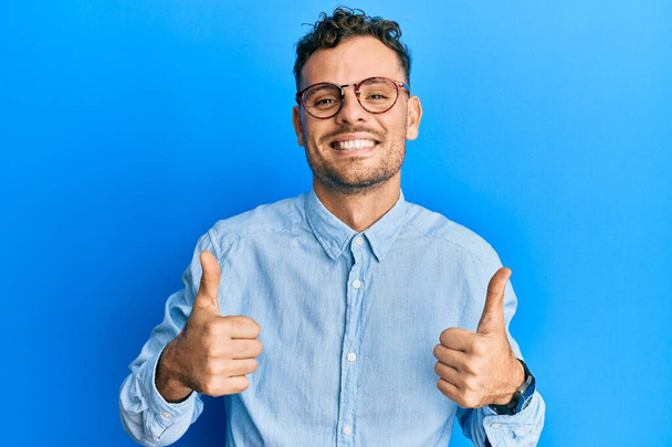 Mladý hispánec v neformálních šatech a brýlích s nápisem "úspěch" dělá pozitivní gesto rukou, palce nahoru, usmívá se a je šťastný. veselý výraz a gesto vítěze.  - Fotografie, Obrázek