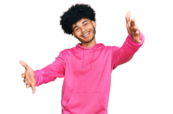Ein junger afrikanisch-amerikanischer Mann mit Afro-Haaren trägt ein lässiges rosafarbenes Sweatshirt und blickt lächelnd mit offenen Armen in die Kamera, um sich zu umarmen. fröhlicher Ausdruck, der das Glück umarmt.  - Foto, Bild