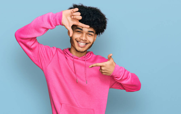 Jonge Afro-Amerikaanse man met afrohaar in casual roze sweatshirt glimlachend makend frame met handen en vingers met gelukkig gezicht. creativiteit en fotografie.  - Foto, afbeelding