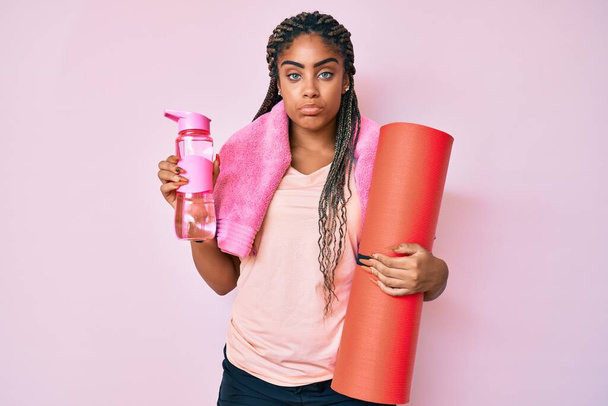 Jonge Afro-Amerikaanse sportieve vrouw met vlechten houdt yoga mat depressief en zorgen te maken voor verdriet, huilen boos en bang. trieste uitdrukking.  - Foto, afbeelding