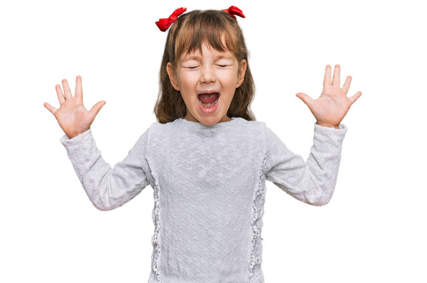 Pieni valkoihoinen tyttö lapsi yllään rento vaatteita juhlii hullu ja hullu menestystä kädet kohotetaan ja suljetut silmät huutaa innoissaan. voittajan käsite  - Valokuva, kuva
