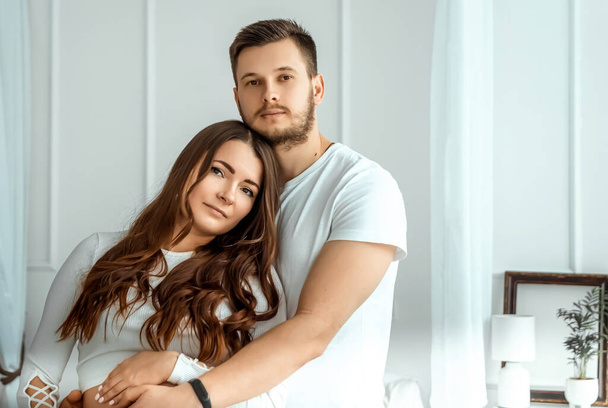 Eine Schwangere und ihr Mann stehen in einem hellen Schlafzimmer. Schöner Bauch einer jungen attraktiven Schwangeren. Familie, Ehe, Geburtenkonzept - Foto, Bild