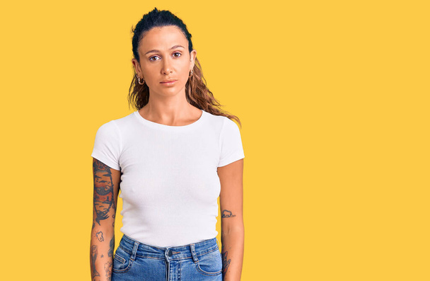 Молодая латиноамериканка с татуировкой в повседневной белой футболке, расслабленная с серьезным выражением лица. простой и естественный взгляд на камеру.  - Фото, изображение