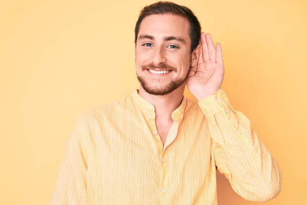 Mladý pohledný muž v neformálních šatech, usmívající se rukou za uchem, naslouchající drbům a drbům. koncepce hluchoty.  - Fotografie, Obrázek