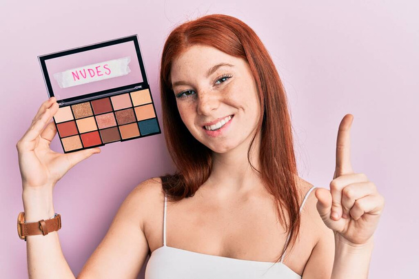Young red head girl holding blush palette of nudes colors smiling with an idea or question pointing finger with happy face, number one  - Photo, Image