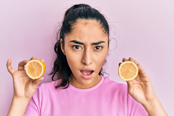 Hispanic teenager girl with dental braces holding lemon in shock face, looking skeptical and sarcastic, surprised with open mouth  - Foto, imagen