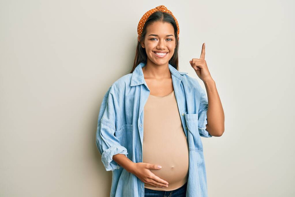 Mooie Spaanse vrouw verwacht een baby, raakt zwanger buik met een grote glimlach op het gezicht, wijzend met de hand vinger naar de zijkant kijkend naar de camera.  - Foto, afbeelding