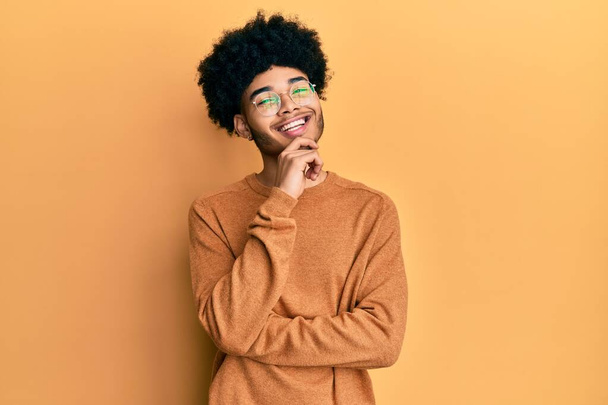 Jovem americano africano com cabelo afro vestindo camisola de inverno casual olhando confiante para a câmera com sorriso com braços cruzados e mão levantada no queixo. pensamento positivo.  - Foto, Imagem