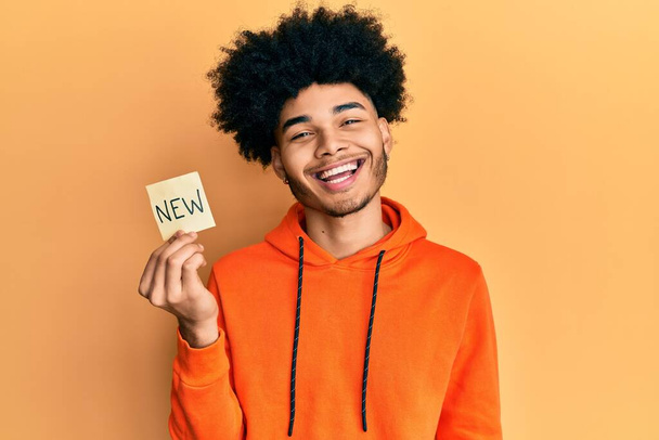 Молодий афроамериканець з волоссям афроамериканця, який тримає нове слово на папері, виглядає позитивним і щасливим, і усміхається з упевненою посмішкою, що показує зуби.  - Фото, зображення
