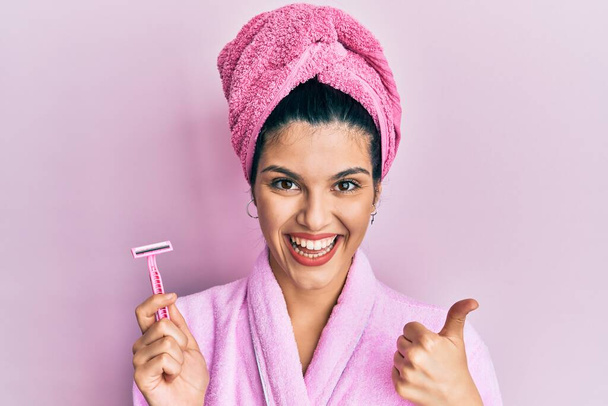 Молодая латиноамериканка в банном халате с бритвой, улыбающаяся счастливая и позитивная, с большим пальцем вверх, отлично справляющаяся и одобряющая табличка  - Фото, изображение