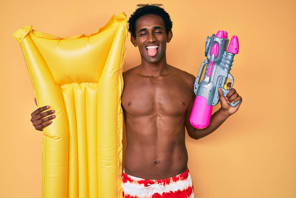 African handsome man wearing swimsuit and holding summer float sticking tongue out happy with funny expression.  - Foto, afbeelding
