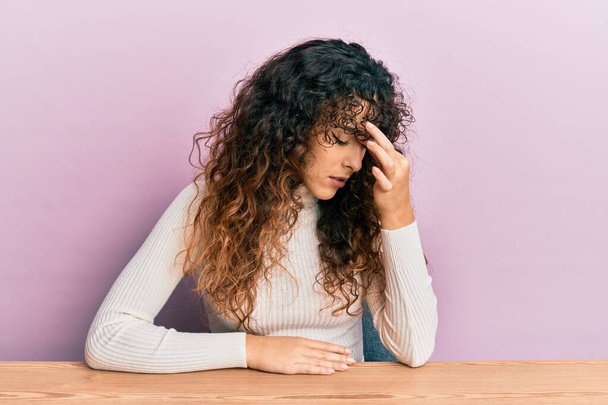 Ein junges hispanisches Mädchen in lässiger Kleidung sitzt auf dem Tisch und reibt sich müde Nase und Augen und spürt Müdigkeit und Kopfschmerzen. Konzept für Stress und Frustration.  - Foto, Bild