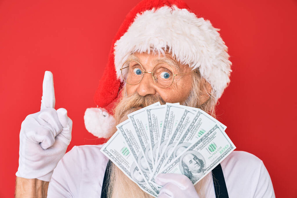 Old senior man with grey hair and long beard wearing santa claus costume holding dollars smiling with an idea or question pointing finger with happy face, number one  - Foto, Imagen