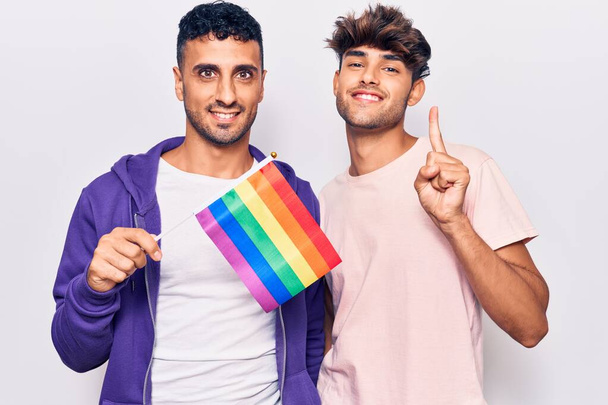Молодая пара геев с радугой lgbtq флаг улыбается с идеей или вопрос указывая пальцем со счастливым лицом, номер один  - Фото, изображение