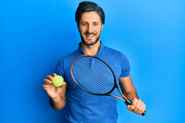 Młody Hiszpan gra w tenisa trzymając rakietę i piłkę uśmiechając się z uśmiechem na twarzy. wykazujące zęby.  - Zdjęcie, obraz