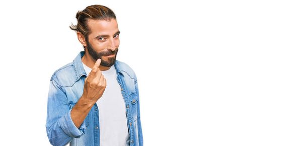 Привлекательный мужчина с длинными волосами и бородой в джинсовой куртке манит сюда жест с рукой приглашая радушно и улыбаясь  - Фото, изображение