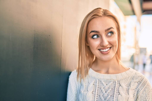 Young blonde girl smiling happy leaning on the wall at the city. - Photo, Image
